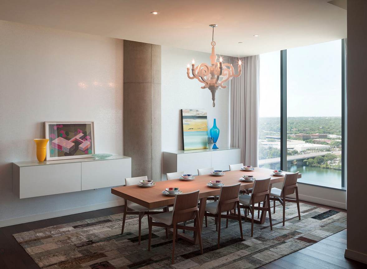 Image depicts a modern dining room with a minimalist style. The layout features a rectangular wooden dining table with eight matching chairs, each with light-colored cushions. The table is set with white plates and bowls, and a few small decorative items. The room has a large floor-to-ceiling window on the right, offering a view of a green landscape and a body of water. The walls are light-colored, with a textured finish. On the left wall, there are two floating white cabinets, each adorned with colorful vases and framed artwork. A contemporary chandelier with a white, ornate design hangs above the dining table. The floor is covered with a patchwork-style rug in neutral tones.