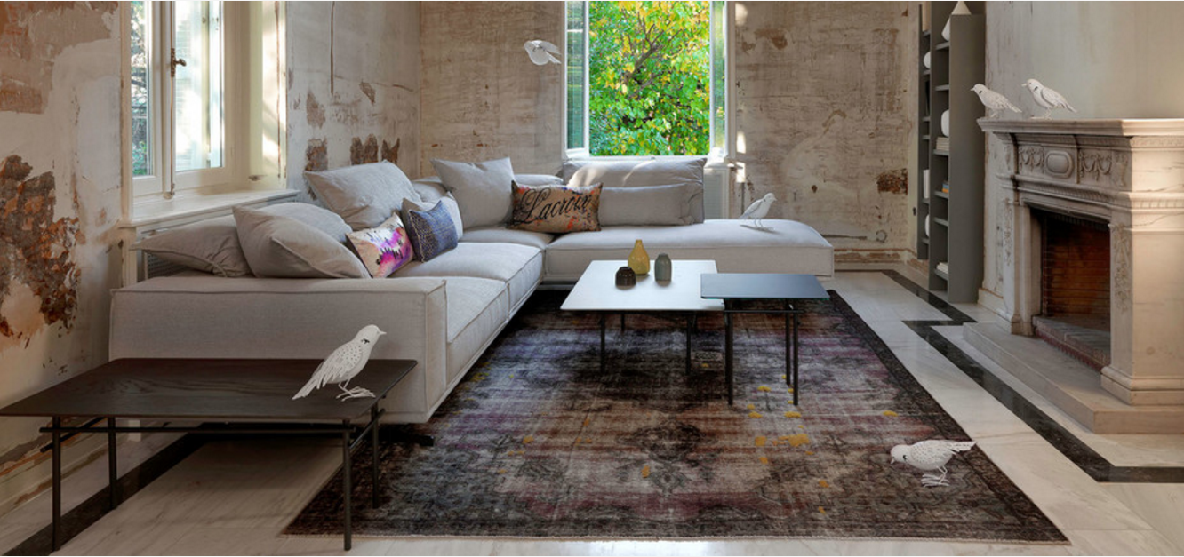 Stylish and modern living room with a rustic touch. The layout features a large, L-shaped light gray sectional sofa adorned with various colorful throw pillows, positioned against a distressed, textured wall. The room has a mix of natural and artificial lighting, with sunlight streaming through a large window that offers a view of lush green foliage outside. The floor is covered with a multicolored, vintage-style rug, and there are two minimalist coffee tables in front of the sofa, one black and one white, each holding small decorative items. A classic, ornate fireplace is situated on the right side of the room, adding a touch of elegance. White bird illustrations are scattered throughout the room, perched on the furniture and window sill, adding a whimsical element to the scene.