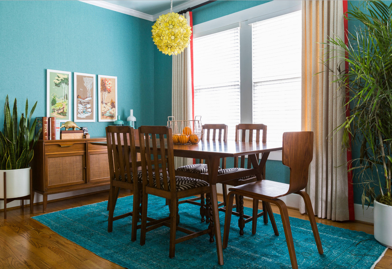 The dining room is furnished with a wooden dining table and five chairs on a 7x10 turquoise area rug. The room that receives daylight is fitted with light-coloured curtains. The walls are painted in a colour that coordinates with the carpet. The floral decorations on either side of the room enhance the overall ambience of the space.