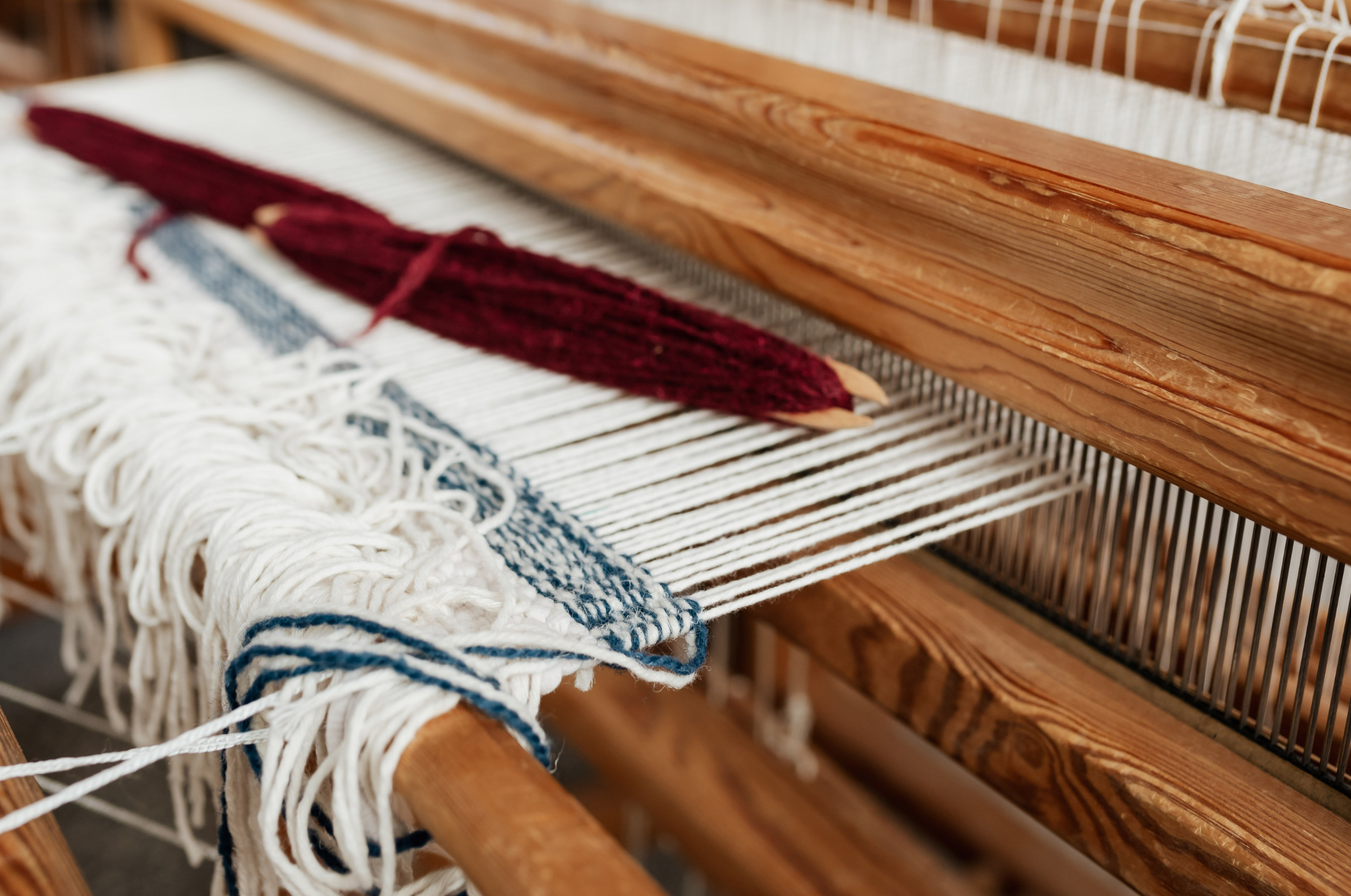 A close-up of a traditional wooden loom with taut white warp threads and a shuttle wrapped in red yarn, illustrating the start of the weaving process.