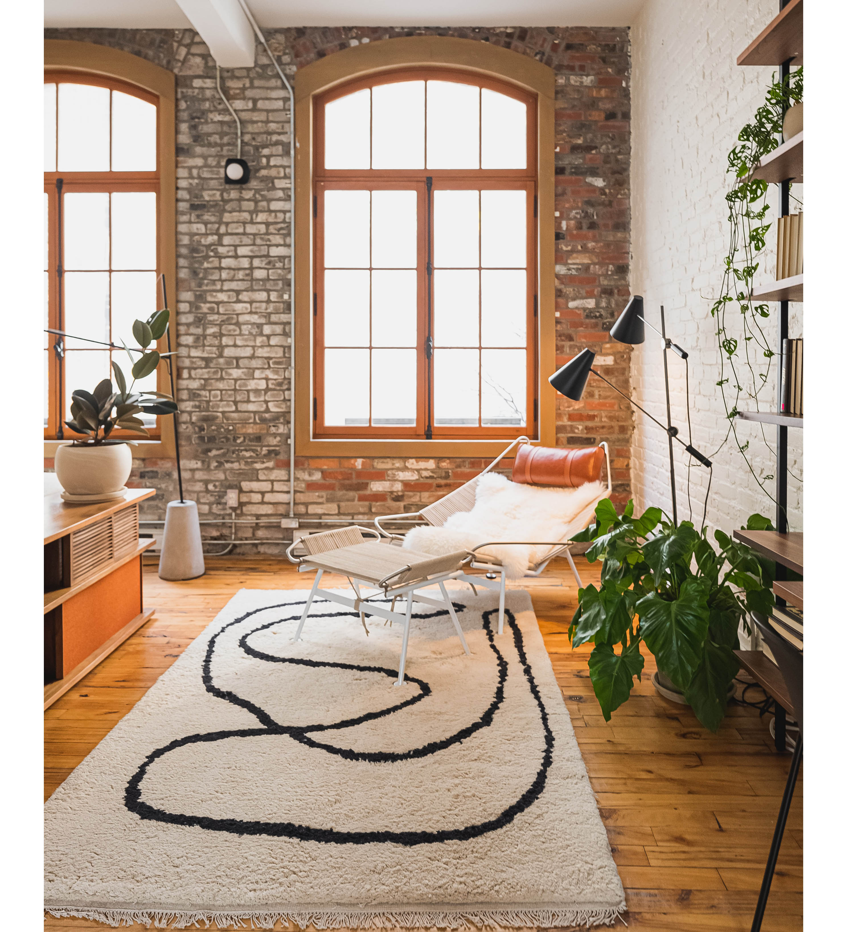 An industrial-style living area decorated with an abstract neutral rug, a lounge chair, and multiple green plants, creating a calm and modern atmosphere.