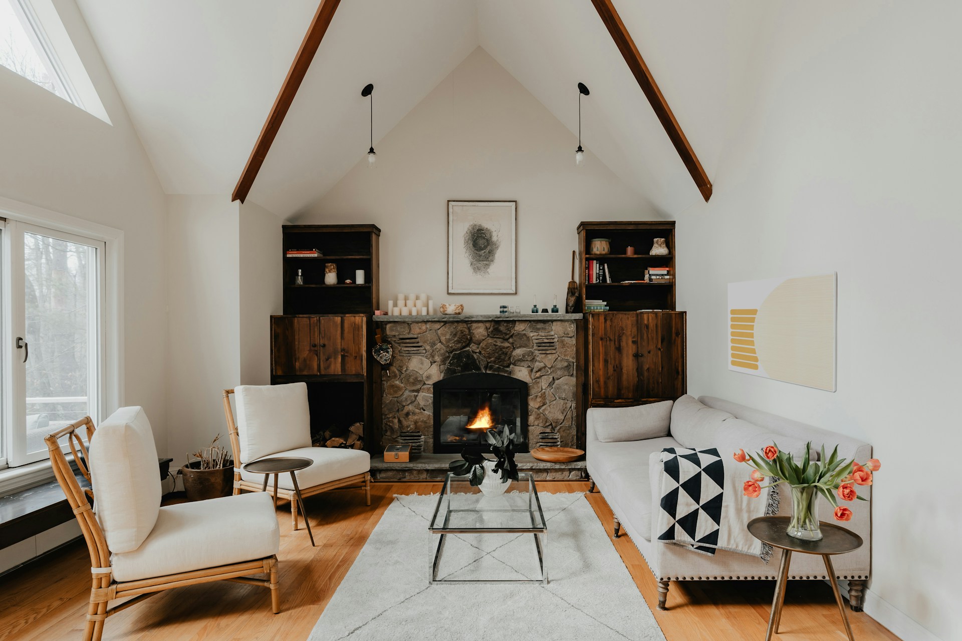 Cozy, modern living room with a high, vaulted ceiling and exposed wooden beams. The room features a stone fireplace at the center, flanked by two dark wooden cabinets with shelves holding books and decorative items. A framed artwork hangs above the fireplace. The seating arrangement includes a light-colored sofa with a geometric-patterned pillow, two cushioned armchairs, and a glass coffee table with a metal frame. A light gray rug covers the wooden floor, and a small side table with a vase of tulips adds a touch of color. Large windows on the left allow natural light to flood the space, enhancing the warm and inviting atmosphere.