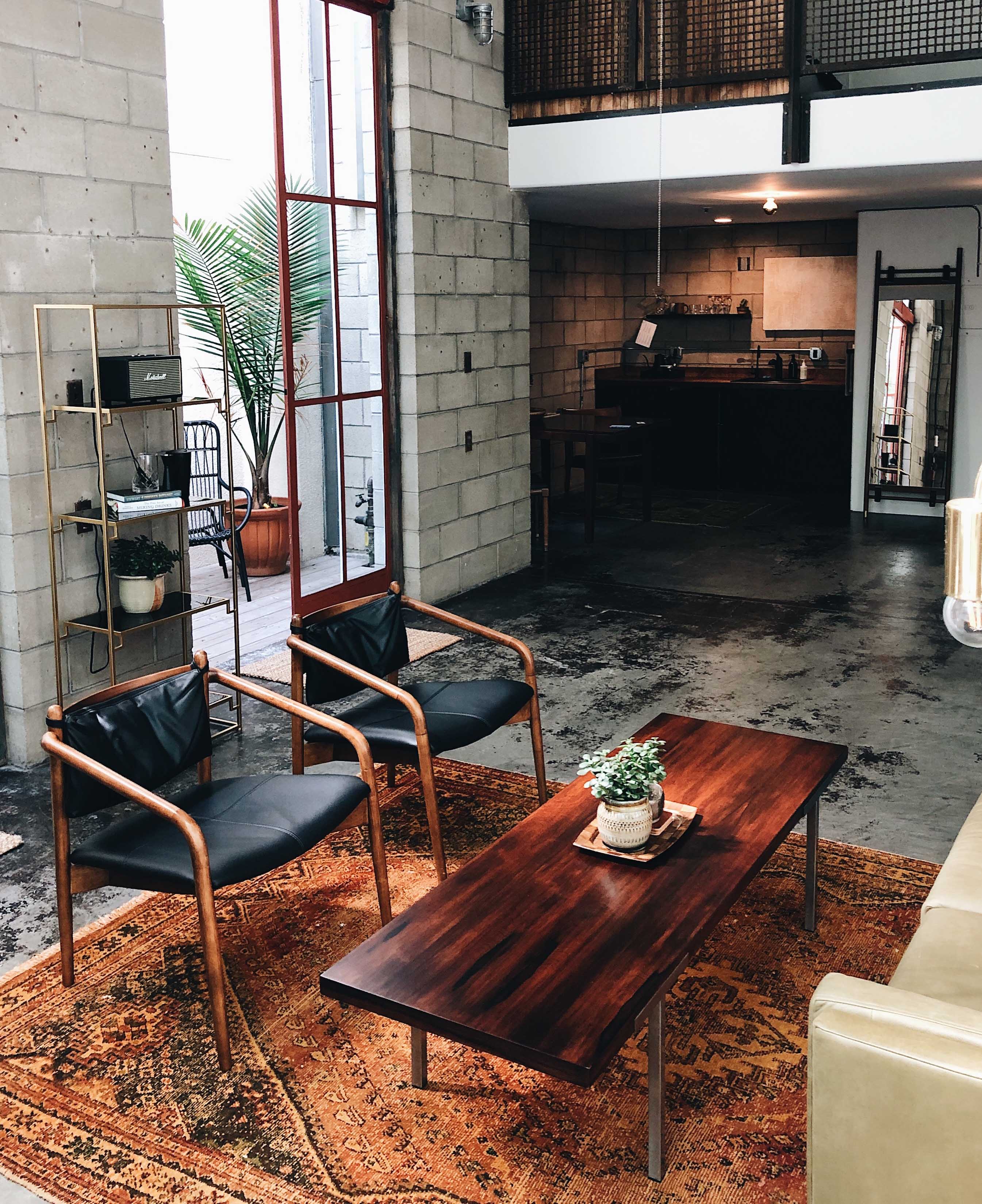 a modern industrial-style living space with an open layout. The room features a concrete block wall and a large window with a metal frame, allowing natural light to flood the area. In the foreground, there is a wooden coffee table with a small potted plant on top, set on a patterned, earthy-toned rug. 