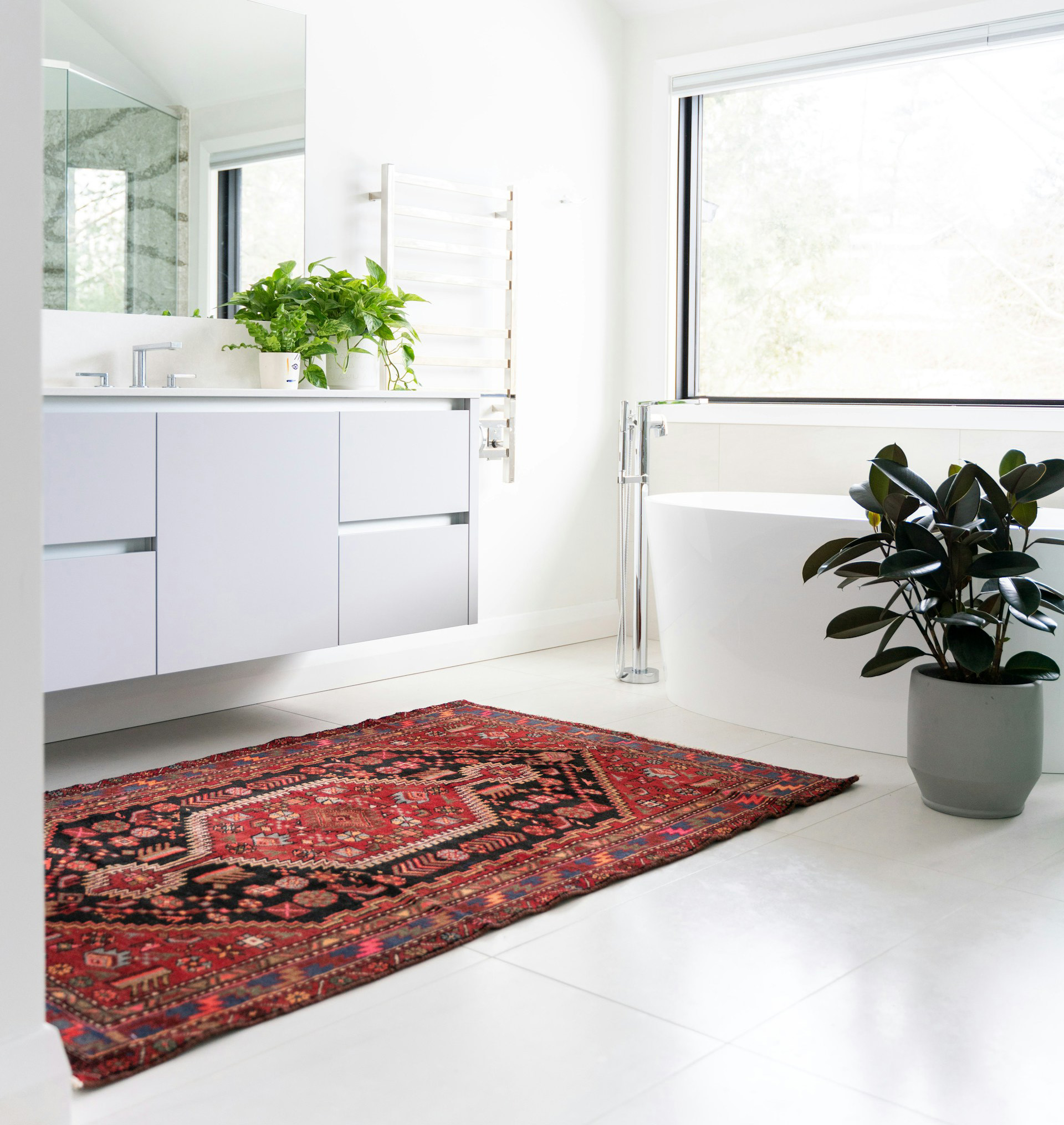 a modern, minimalist bathroom with a bright and airy layout. The room features a large window on the right, allowing natural light to flood the space. A sleek, white vanity with flat-panel cabinets is mounted on the left wall, topped with a white countertop and a rectangular mirror above. A lush green plant sits on the vanity, adding a touch of nature. In the foreground, a vibrant red and black patterned rug with intricate geometric designs covers the white tile floor. A freestanding white bathtub is positioned near the window, accompanied by a tall, leafy plant in a gray pot. The overall color scheme is predominantly white, with accents of green and red, creating a clean and inviting atmosphere.