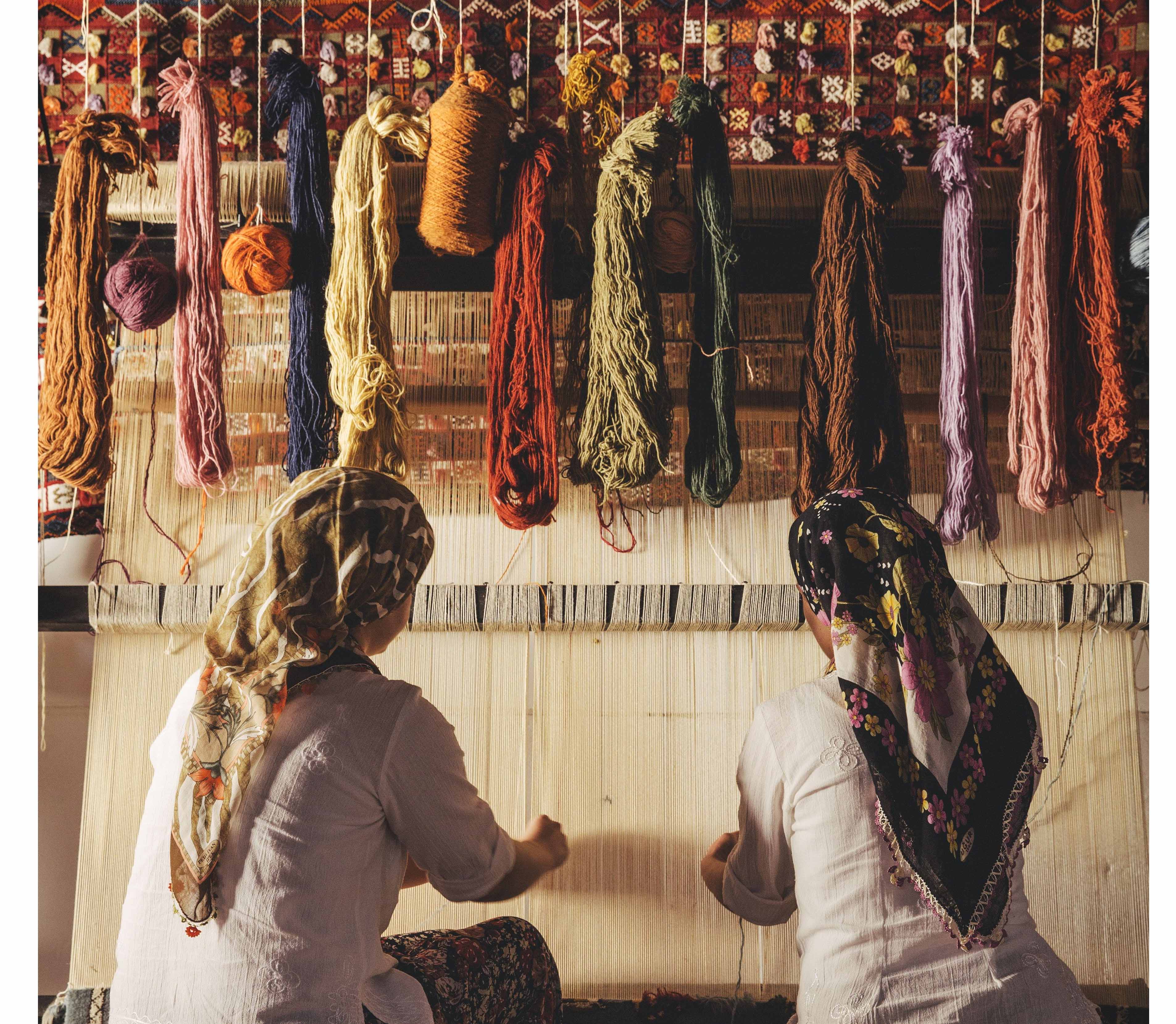 The start of a hand-knotted rug's journey, showing two weavers using a traditional loom to transform colorful yarns into a beautiful, lasting piece of art.