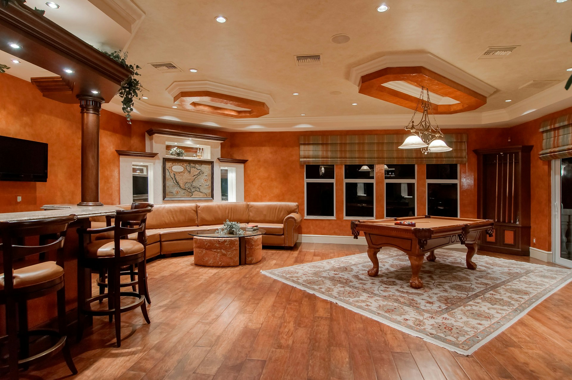 An interior environment characterised by the predominant presence of the colour orange. The interior design features an orange wall colour scheme, a beige ceiling, and leather seating. The lighting is designed to be unobtrusive and enhances the spacious feel of the environment. Positioned beneath the billiard table is a 10 x 12 hand-woven beige base with a hand-woven oriental motif Turkish carpet, featuring a predominantly red and orange colour scheme. The space is designed with relaxation and rest in mind and has a welcoming ambience.