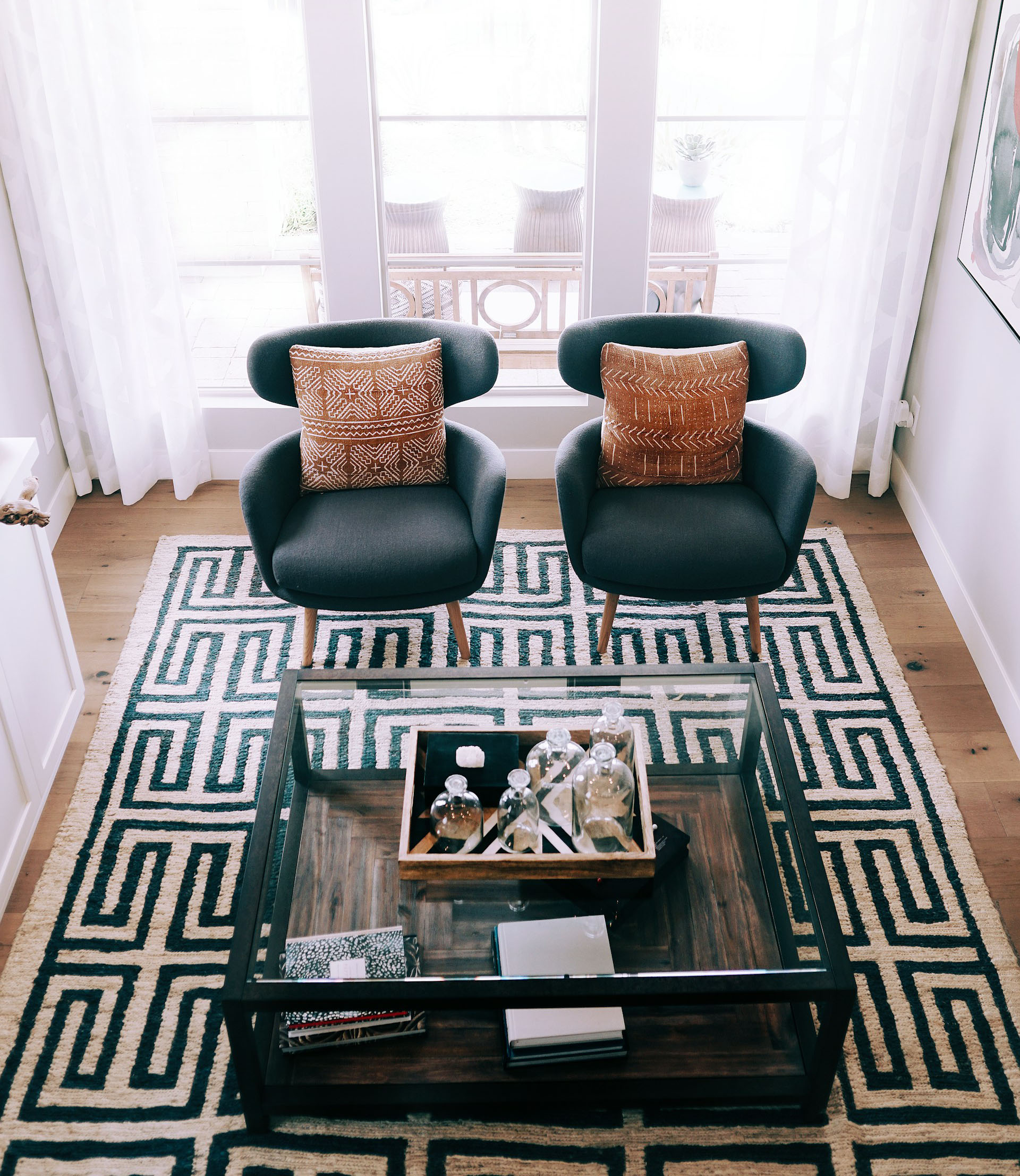 The room is anchored by a substantial hand-woven Turkish rug, showcasing a serene geometric pattern in beige and black. The rug, measuring 10 by 12 feet, exudes a sense of tranquility. The seating area is enhanced by the addition of dark armchairs and vibrant cushions. The soft patterns and tones of the rug are enhanced by the warmth of natural light. The ambience is designed to create a harmonious environment that symbolises comfort and peace.
