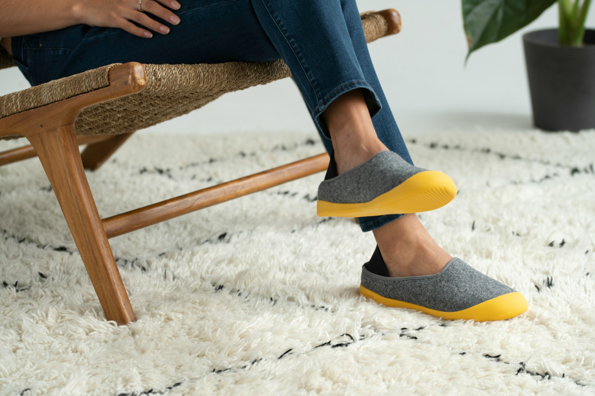 The photograph displays the feet of an individual seated on a white pinstriped, Beni Ourain carpet and only shows the feet of a modern-style wooden armchair with a flower vase and a flower positioned at the back.