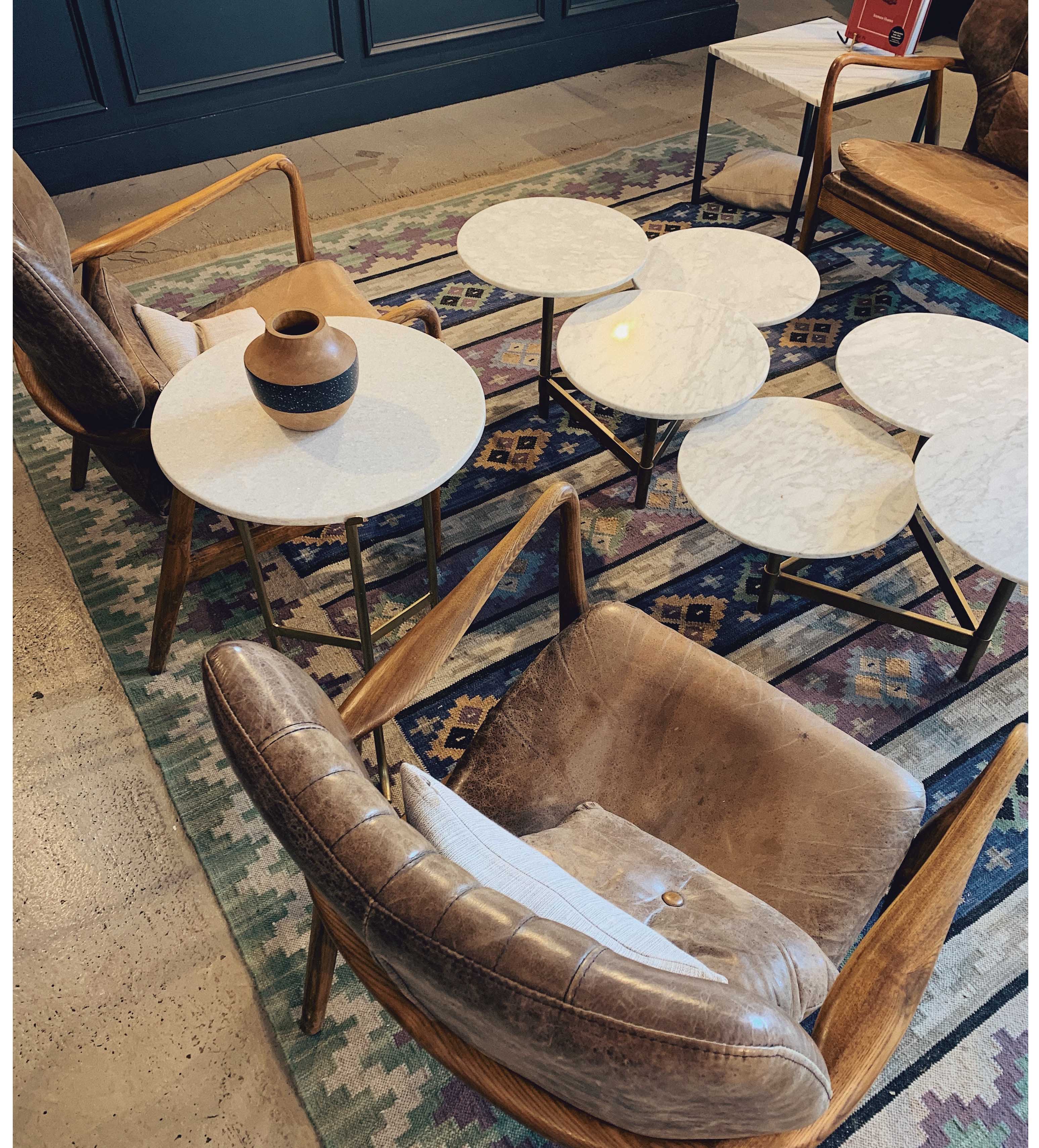 Overhead view of a modern living area featuring a multicolored flatweave kilim rug with geometric patterns in blues, purples, and creams, anchoring two leather armchairs and a cluster of marble-top side tables.