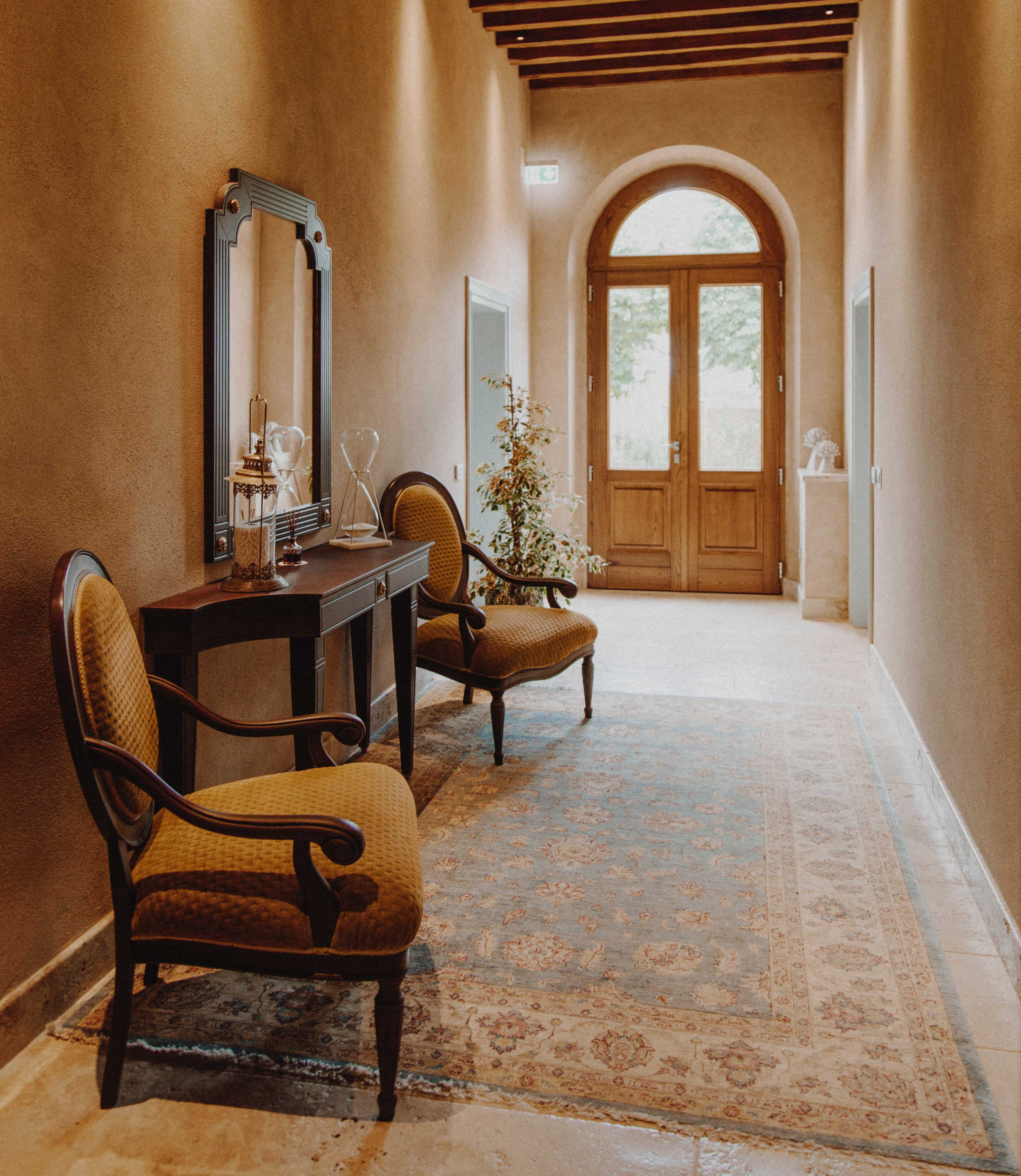 a stylish and elegant hallway with a classic design. The layout features a long, narrow corridor with a high ceiling and exposed wooden beams. The walls are painted in a warm, neutral tone, complementing the polished stone floor. At the end of the hallway, a large arched wooden door with glass panels allows natural light to filter in, illuminating the space. On the left side, a dark wooden console table is adorned with decorative glassware and a mirror with a black frame. Two upholstered chairs with curved wooden arms and legs are positioned symmetrically on either side of the table. A patterned rug in muted colors runs along the length of the hallway, adding texture and warmth to the setting. A small potted plant is placed near the door, enhancing the inviting atmosphere.