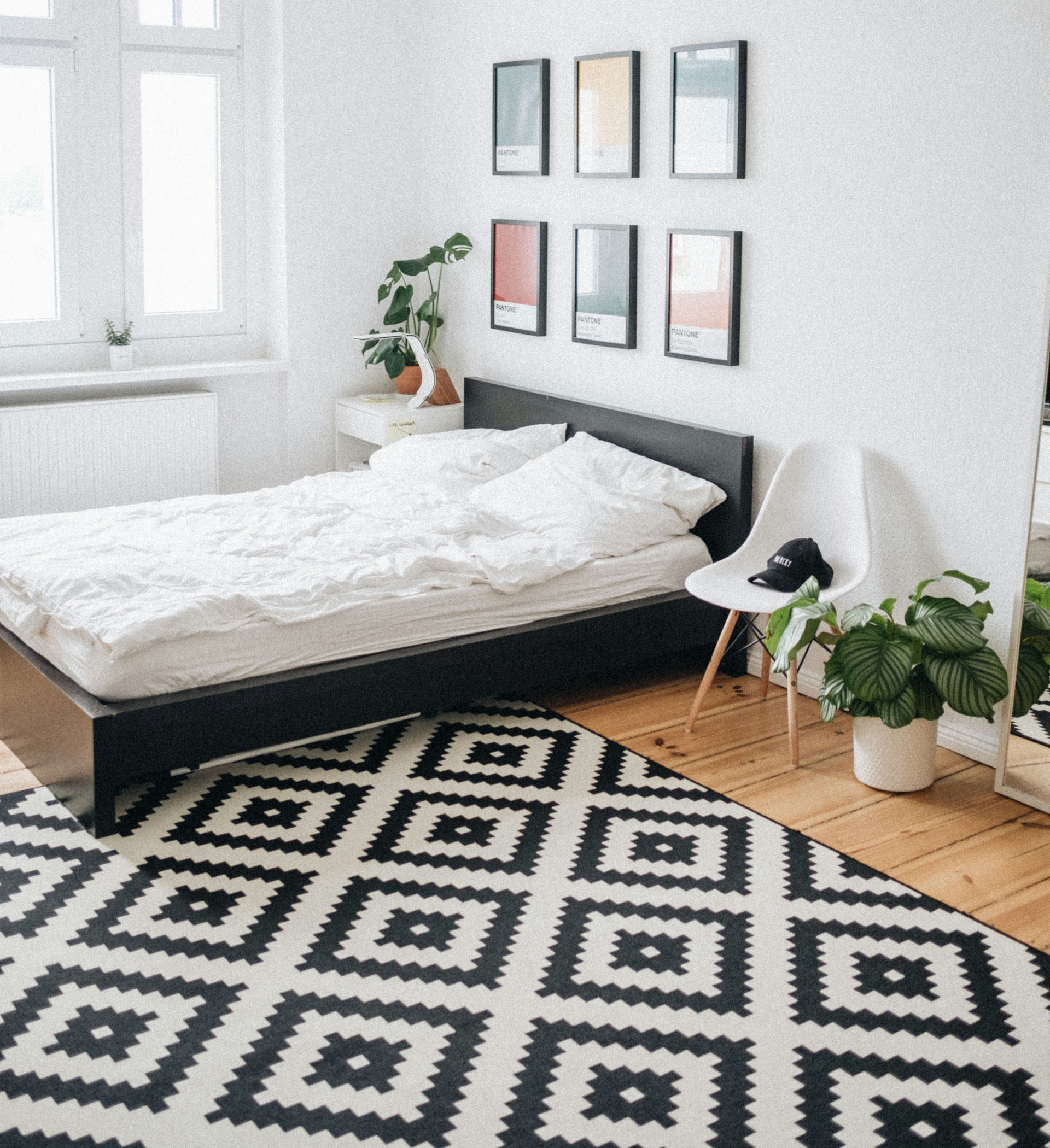 The bedroom is designed in a contemporary, minimalist style with a clean and uncluttered layout. The room is furnished with a low-profile black bed frame and white bed linen, positioned against a white wall. The wall above the bed features a grid of six framed prints, each with a different colour and design. To the left of the bed, a potted plant is positioned on a small white nightstand. A white chair with a black headboard is positioned adjacent to a large potted plant with broad green leaves. The floor is covered with an 5x8 area rug featuring a black and white geometric pattern. The large window on the left side allows natural light to flood the room, creating a bright and airy atmosphere.