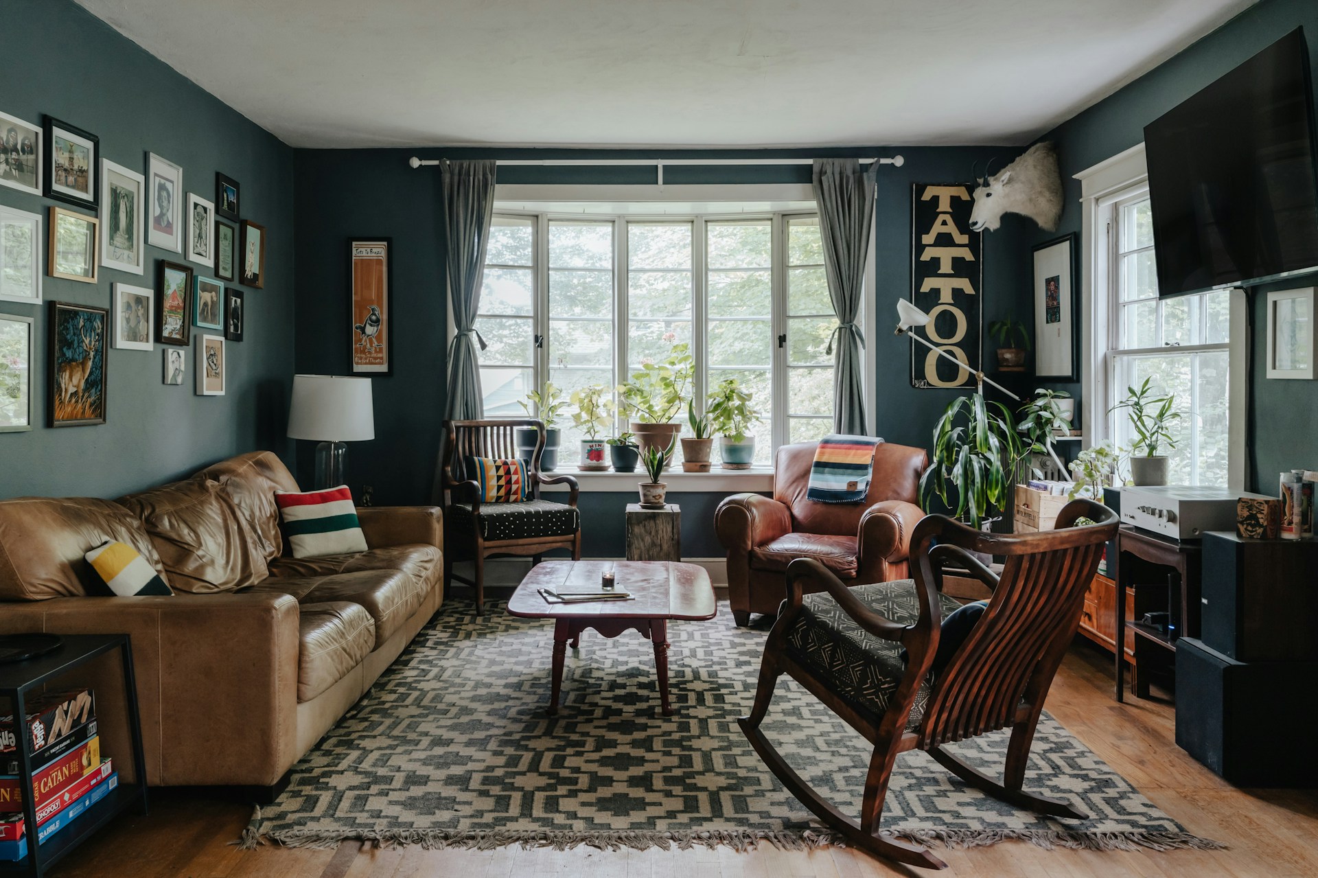 a stylish and eclectic living room with a cozy and inviting atmosphere. The layout features a large window with white frames, allowing natural light to illuminate the space. The walls are painted in a dark teal color, adorned with an array of framed pictures and artwork, adding a personal touch. A brown leather sofa and two armchairs, one in brown leather and the other in dark wood with a cushioned seat, are arranged around a wooden coffee table on a geometric-patterned rug. 