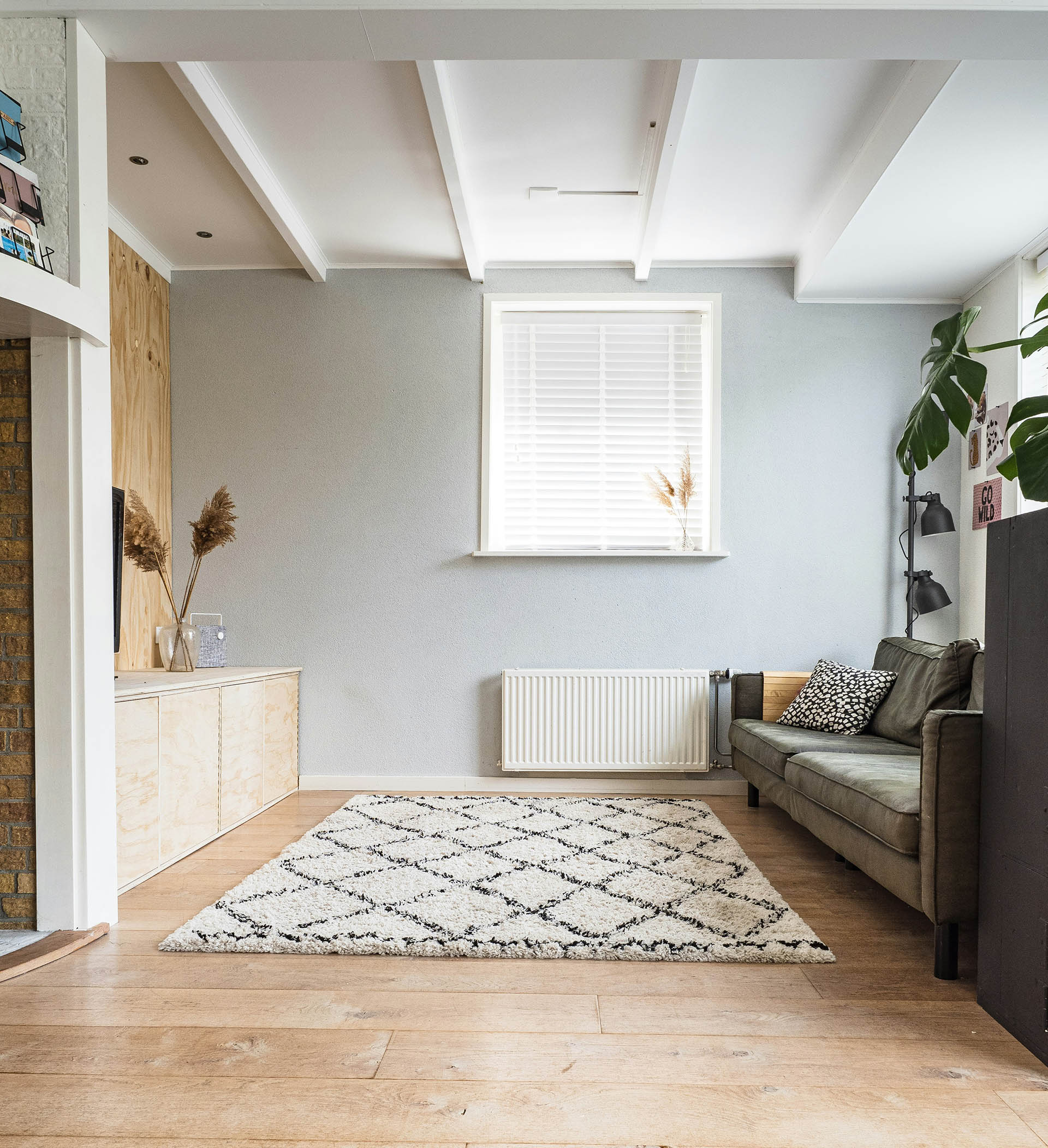 The striking white Moroccan rug with an open weave creates a light and airy effect. On the left, the green sofa and TV unit provide a comfortable seating area. The room is elegantly decorated and illuminated by soft, natural light from a lamp.