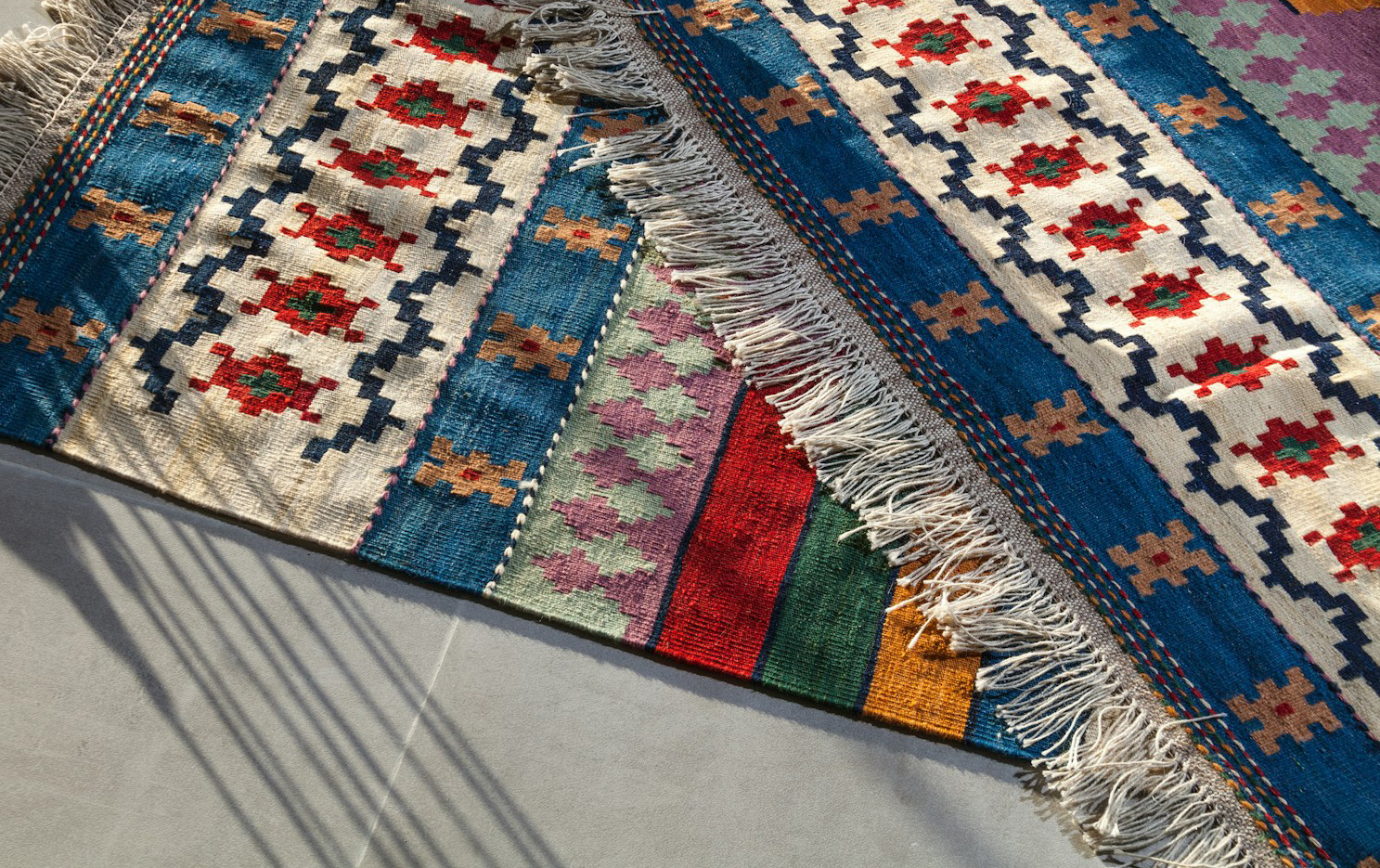 Close-up view of a vibrant traditional flatweave kilim rug with geometric patterns in blue, white, red, and green, featuring visible fringe and hand-loomed texture.
