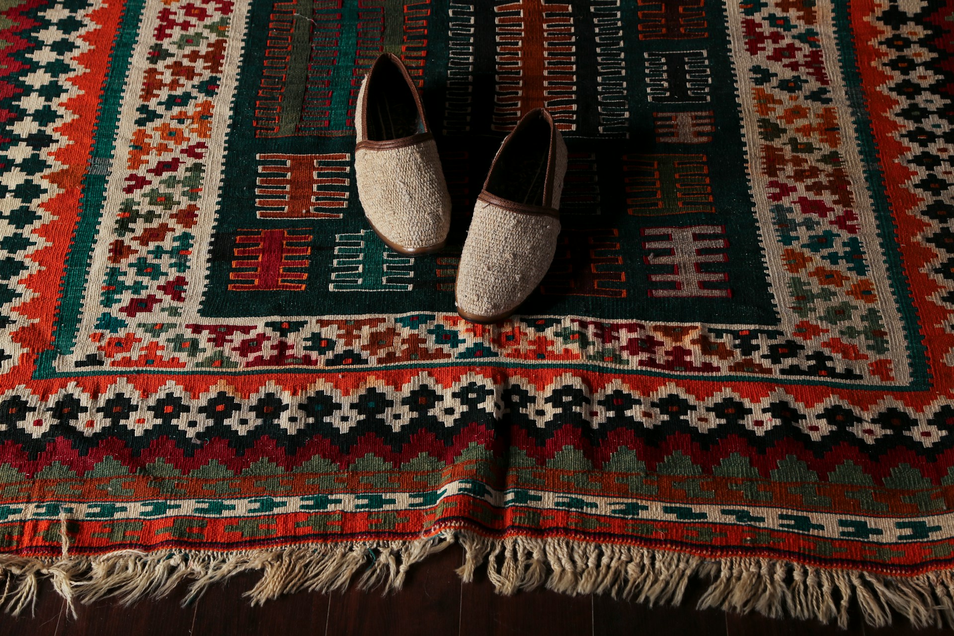 Overhead shot of a vibrant Turkish handwoven flatweave kilim rug with intricate geometric patterns in green, orange, red, and cream, featuring visible fringe and a pair of traditional slippers on top.