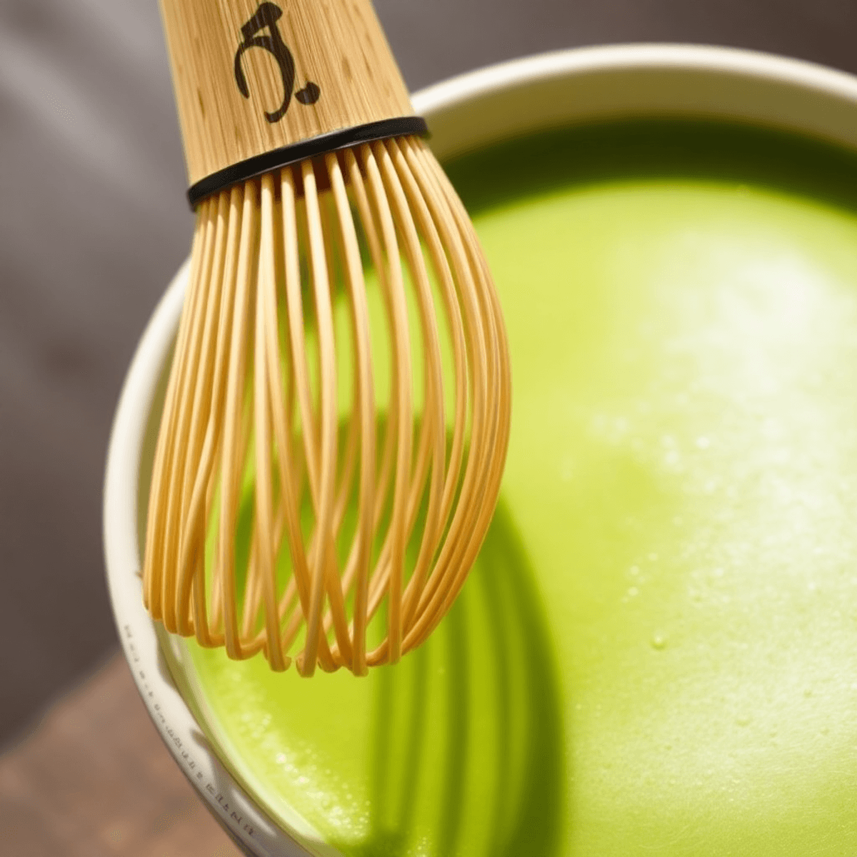 how to whisk matcha without a matcha whisk Close-up of a bamboo matcha whisk beside a vibrant green bowl of frothy matcha tea with soft natural lighting, highlighting the delicate bamboo and creamy foam.