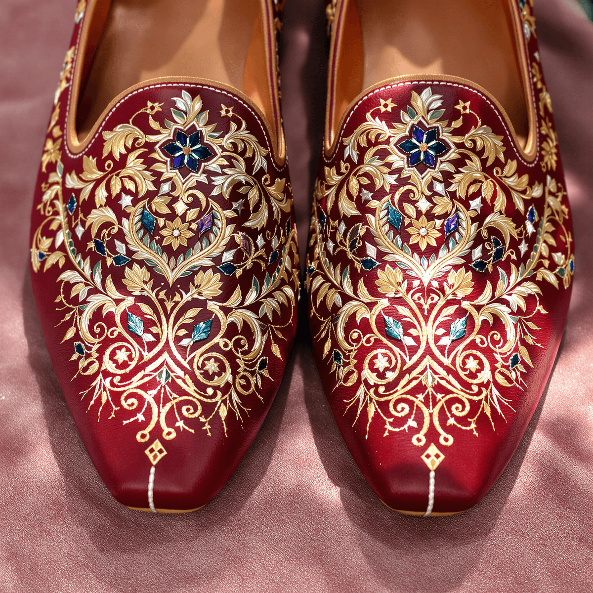 Close-up of elegant maroon leather khussa shoes with intricate hand-stitched embroidery on textured fabric, highlighted by soft natural lighting.