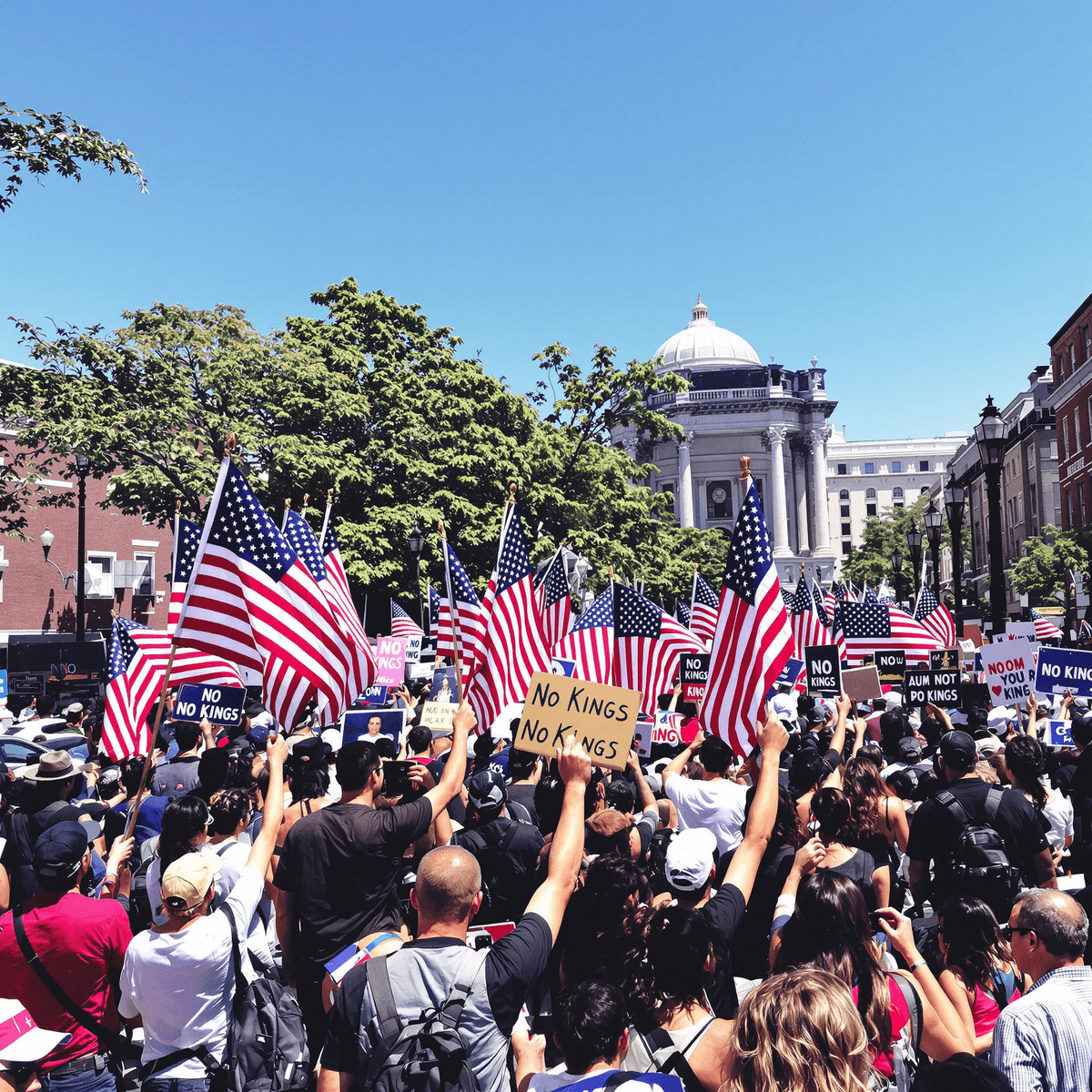 A large crowd of protesters holding American flags and "No Kings" signs in a vibrant urban square under a clear blue sky.